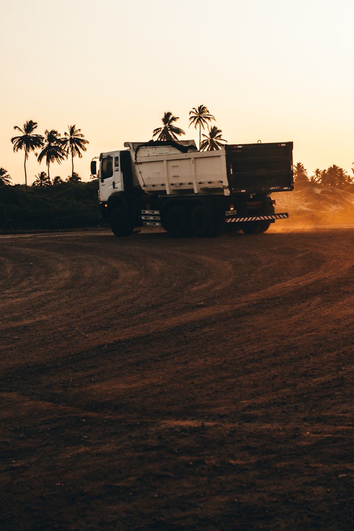 A large dump truck moves across a dusty landscape at sunset with palm trees in the background.